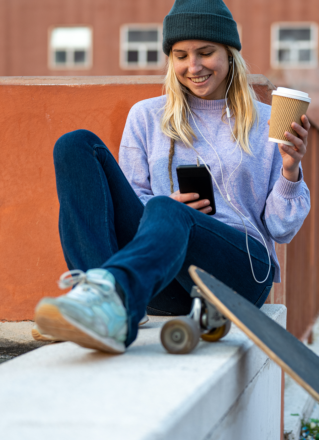 a young woman with sits with headphones, her skateboard, a coffee, and her phone